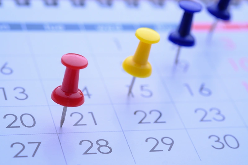 Gros plan sur un calendrier papier avec des punaises color&eacute;es plant&eacute;es sur plusieurs dates, illustrant des &eacute;ch&eacute;ances importantes.
