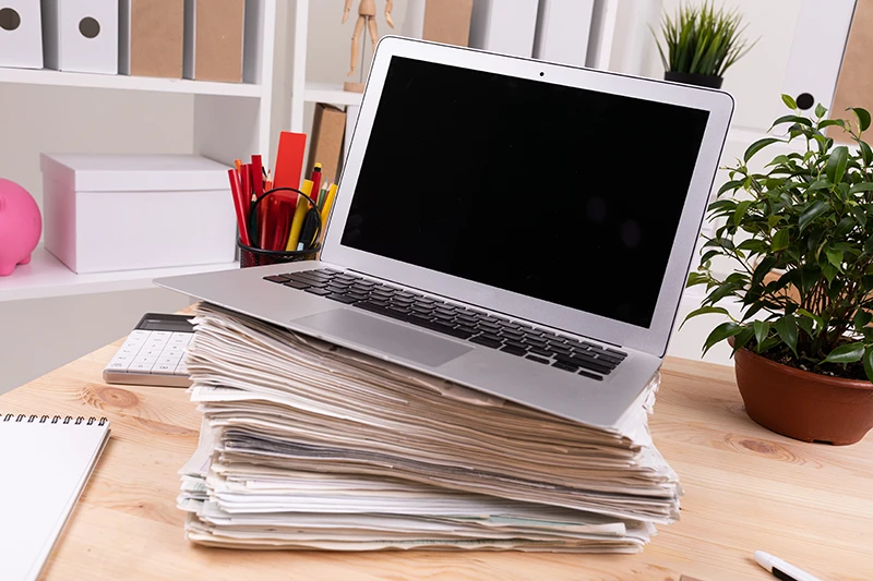 Ordinateur portable pos&eacute; sur une pile de documents papier dans un bureau, illustrant la surcharge administrative avant la d&eacute;mat&eacute;rialisation.