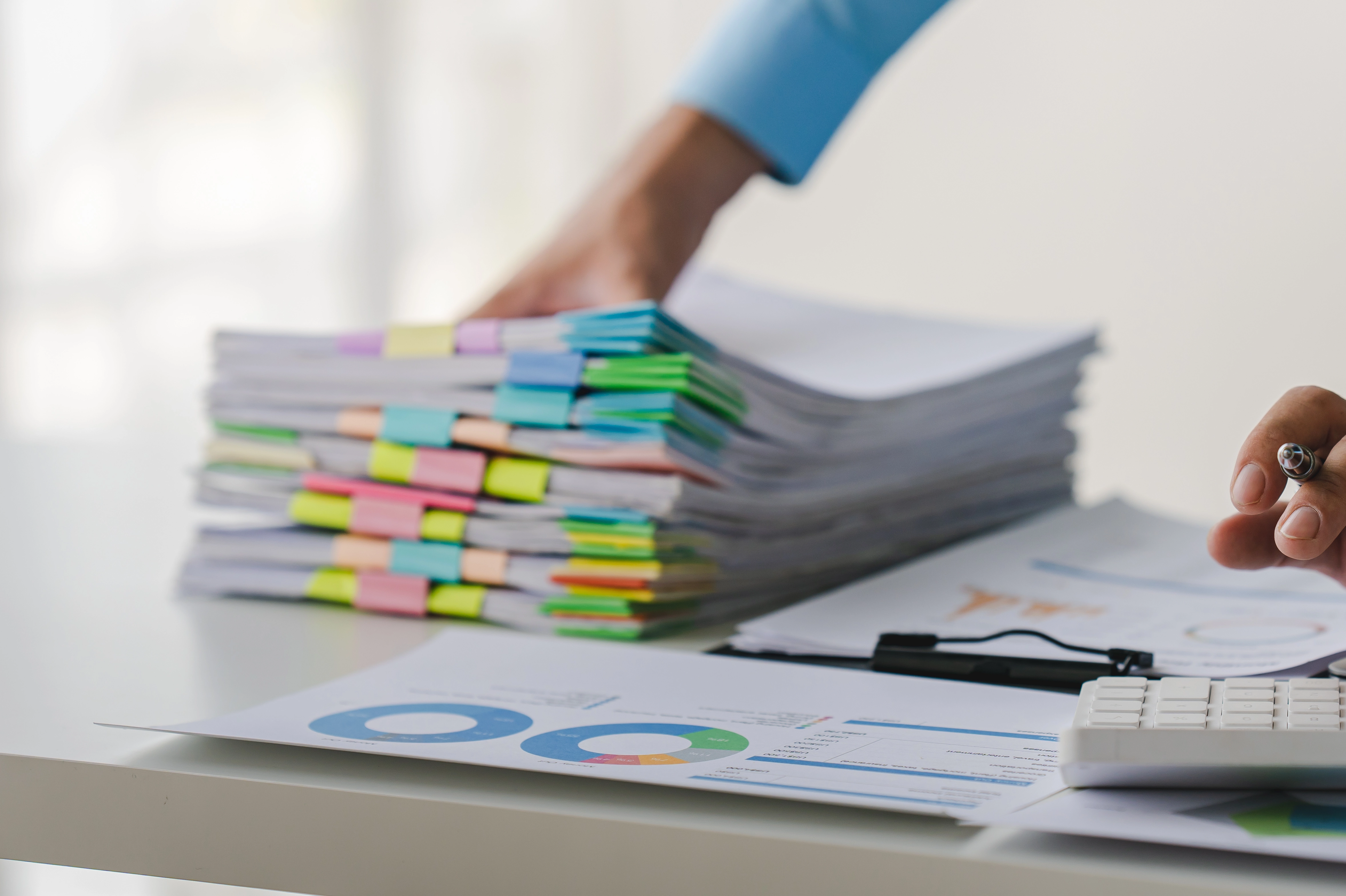 Main d&rsquo;un professionnel manipulant une pile de dossiers class&eacute;s par couleurs, sur un bureau d&rsquo;entreprise avec documents et calculatrice